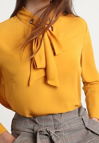 Gelbe Bluse mit einem Schleifen-Ausschnitt und glattem Stoff. Kombiniert mit einem karierten, grauen, hochgeschnittenen Rock mit einem gebundenen Detail.