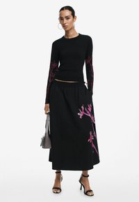 Top nero a maniche lunghe con ricami floreali rosa sulle maniche, abbinato a una gonna lunga nera con un design floreale rosa simile.