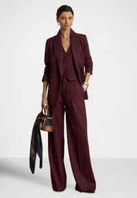 Bordo spalvos trijų dalių kostiumas su pritaikytu švarku, liemene ir plačiomis kelnėmis; tekstūruota medžiaga, mygtukų detalės ir aksesuarų krepšelis su skarele.