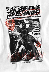 Weißes T-Shirt mit einem grafischen Design in Schwarz und Rot. Das Bild zeigt ein Wesen mit einem blumigen Kopf, Flammen und Text über mysteriöse Sichtungen.