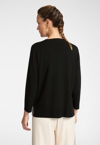 Mujer con cabello trenzado, usando aretes de oro en forma de aro, una blusa negra de mangas largas y pantalones blancos, de espaldas frente a un fondo liso.