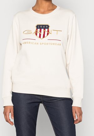 Kremowa bluza z kapturem z czerwono-biało-niebieską tarczą i napisem "GANT 1949 American Sportswear", noszona z ciemnymi dżinsami, widok z przodu przycięty na wysokości szyi i ud.