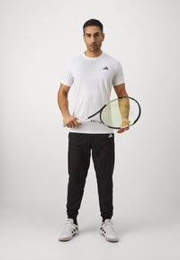 Valge sportlik T-särk tekstuurse mustriga, must logo; mustad püksid; hoiab tennisereketit, millel on kollased nöörid; valged tenis kingad.