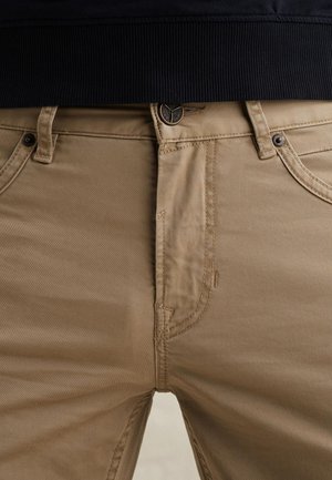 Nahaufnahme von beigen Hosen mit Gürtelschlaufen, Knopf und Taschen, kombiniert mit einem dunkel gerippten Oberteil.