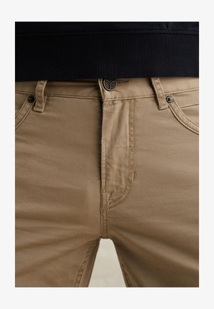 Nahaufnahme von beigen Hosen mit Gürtelschlaufen, Knopf und Taschen, kombiniert mit einem dunkel gerippten Oberteil.