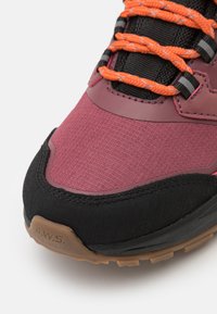 Tursko for hiking med en maroon overdel, teksturert stoff, svarte detaljer og oransje snørebånd. Har forsterket tåhette og logo på hælen.