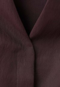 Bordowy materiał o gładkiej fakturze i subtelnym połysku, złożony przy kołnierzu w formie fałdy i z czystym, przeszytym szwem.