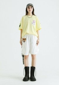 Camiseta amarilla de gran tamaño con gráfico de dibujos animados, pantalones cortos cargo blancos con parche de león y botas negras con cordones. Atuendo urbano casual.