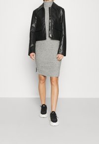 Donna che indossa un vestito a collo alto grigio in maglia, una giacca in pelle nera tagliata e Sneakers in bianco e nero, che si trova di fronte a uno sfondo bianco semplice.