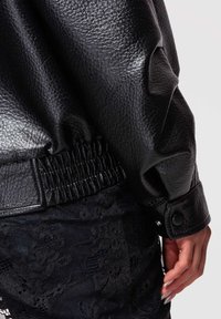 Primer plano de una mano descansando cerca de la manga de una chaqueta de cuero texturizado negra con botón a presión y una prenda de encaje negro debajo.