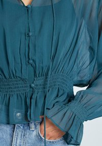 Blusa leggera color turchese con bottoni davanti, vita arricciata e maniche a volant. Realizzata in tessuto trasparente e testurizzato.