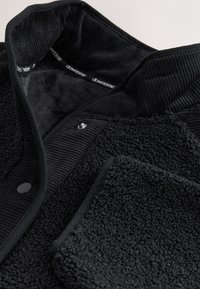 Veste en polaire noire avec un extérieur texturé, un col en velours côtelé, une fermeture à pression et une doublure intérieure douce. Comprend une poche latérale.
