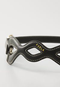 Cintura in pelle nera con un design ondulato e elementi ritagliati, hardware dorato e cuciture bianche a contrasto. Logo "Furla" in giallo.