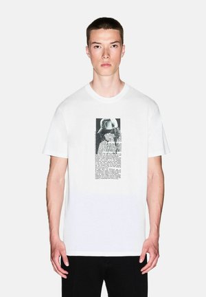 Giovane uomo con capelli corti che si trova frontalmente, indossando una maglietta bianca con una grafica in bianco e nero e testo centrato sul petto.