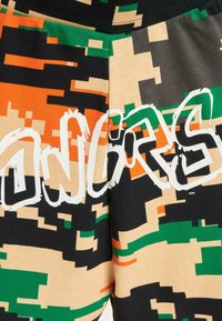 Multifarvede joggers har et pixeliseret camouflage mønster i grøn, orange, sort og beige, med ordet "TILLYKKE" i hvid skrift.