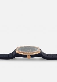 Orologio ottagonale con una lunetta in oro rosa, dettagli in cristallo trasparente, quadrante scuro e cinturino in silicone blu navy. Corona rotonda sul lato.