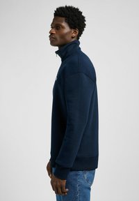 Sweat-shirt zippé bleu marine avec un col montant, des poignets et un ourlet côtelés, fabriqué en tissu doux. La vue de côté montre un design ajusté.