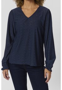 Blouse bleu marine à manches longues avec un col en V, présentant des motifs ajourés texturés et des poignets élastiqués, associée à un jean en denim foncé.