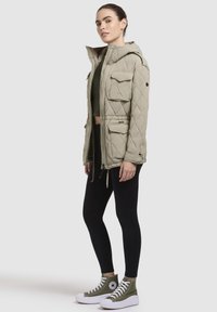 Gequiltede olijfgroene parka-jack met voorkruiszakken, verstelbare capuchon en trekkoord in de taille. Gecombineerd met zwarte leggings en hoge sneakers.