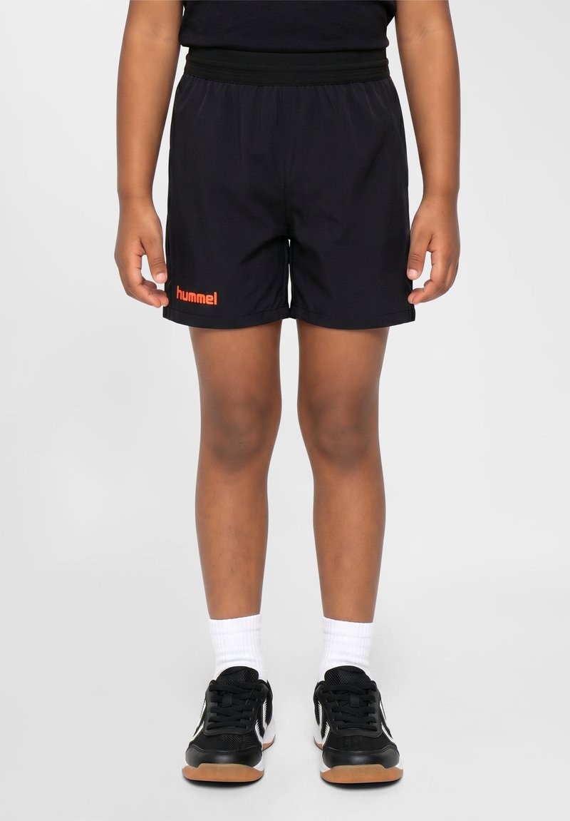 Enfant portant un short de sport noir hummel, des chaussettes blanches montantes et des chaussures de sport noires, debout sur un fond gris clair.