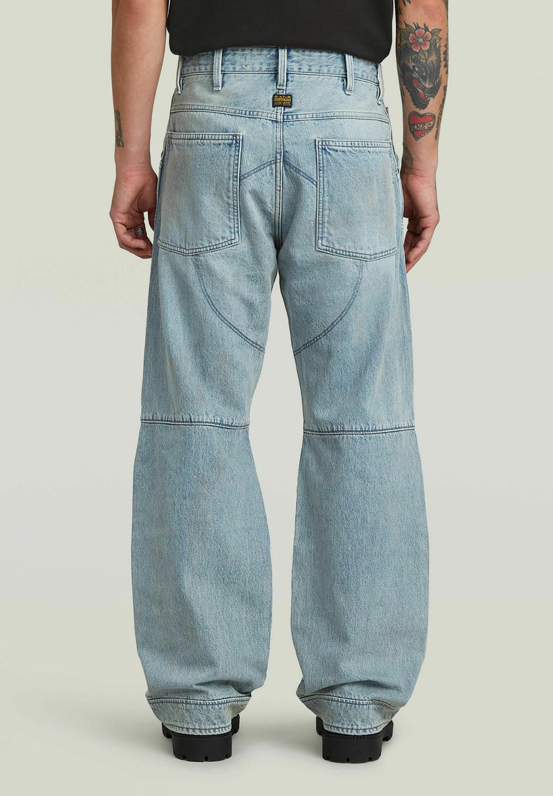 Persona che indossa jeans a gamba larga di colore azzurro chiaro con tasche posteriori, cuciture visibili e scarpe nere, in piedi davanti a uno sfondo semplice.