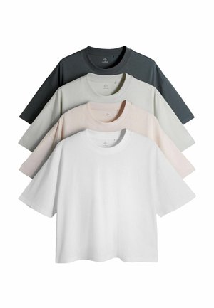 Ung mand stående med hænderne i lommerne iført hvid kortærmet skjorte, mørkeblå vidde jeans og sorte sko mod ensfarvet baggrund.