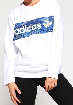 Person iført en hvid Adidas-sweatshirt med en blå mønstret logo og sorte bukser, poserer med den ene hånd ved hoften.