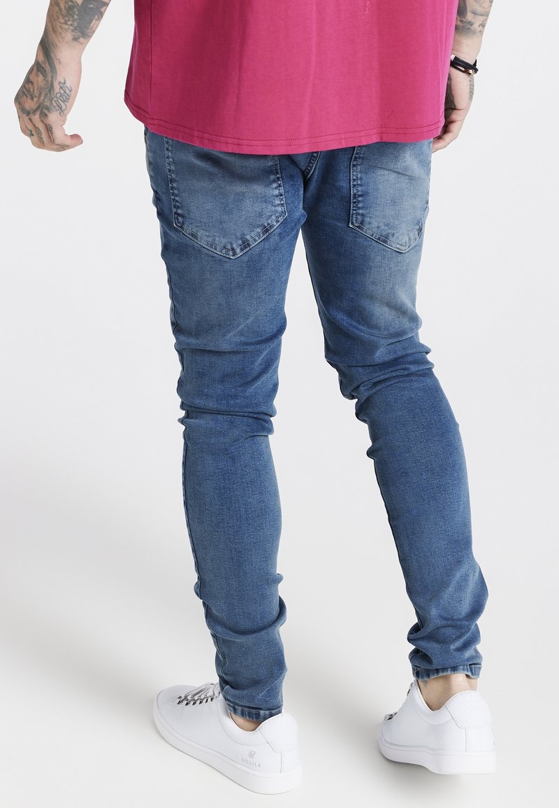 Jeans de denim azul ajustados con un acabado desgastado, con bolsillos de parche, corte slim y combinados con zapatillas blancas.