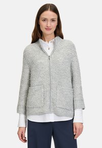 Hellgrauer Strickcardigan mit strukturiertem Muster, Reißverschluss, gerippten Bündchen und zwei vorderen Taschen. Über einem weißen Hemd getragen.
