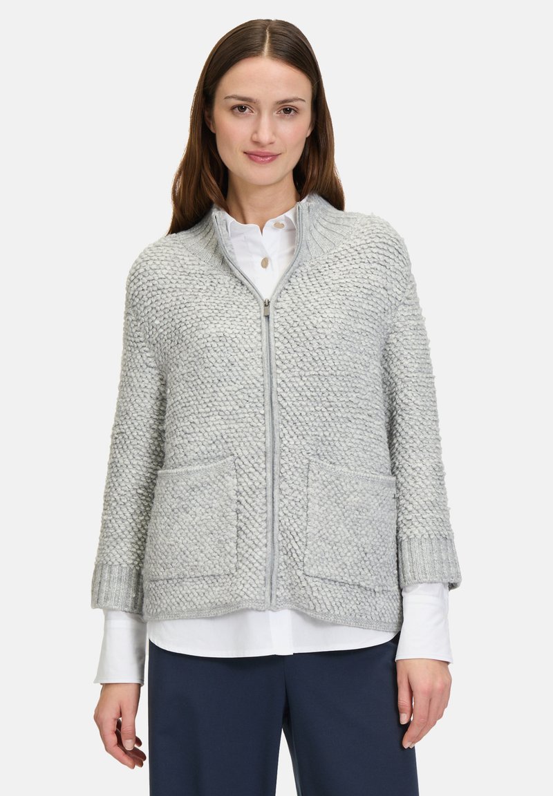 Hellgrauer Strickcardigan mit strukturiertem Muster, Reißverschluss, gerippten Bündchen und zwei vorderen Taschen. Über einem weißen Hemd getragen.