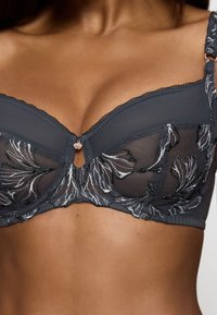 Černá krajková podprsenka s květinovou výšivkou ve stříbrné barvě, s podprsenkovou výztuží, průhlednými panely a centrálním otvorem zdobeným dekoračním charmem.
