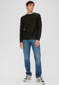 Zwarte langslavende shirt met een ronde hals, gecombineerd met lichtblauwe jeans en chunky sneakers, tegen een effen grijze achtergrond.