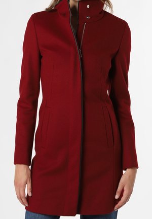 Femme portant un manteau ajusté rouge foncé, longueur genou, avec col montant, manches longues et fermeture éclair et poches avant dissimulées.