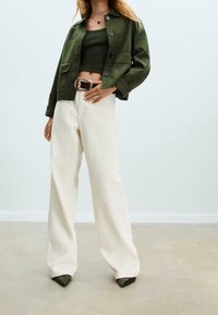 Veste en daim vert avec deux poches avant, associée à un top vert ajusté, un pantalon large couleur crème et des escarpins noirs à bout pointu.