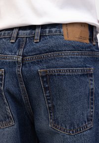 Σκούρο μπλε τζιν με πορτοκαλί ραφές, δερμάτινη ετικέτα με λογότυπο στη μέση και εμφανείς πίσω τσέπες.