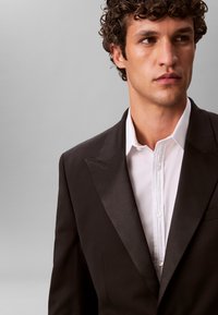 Homme aux cheveux bruns bouclés portant une veste de smoking noire et une chemise blanche, regardant sur le côté sur un fond gris uni.