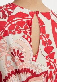 Blouse rouge à motifs avec une ouverture en goutte d'eau à l'arrière, dotée d'un petit bouton rouge. Fabriquée en tissu lisse et léger.