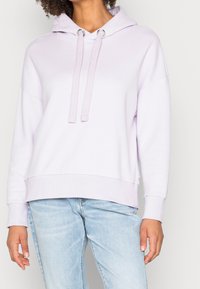 Sudadera con capucha lila con bolsillo frontal tipo canguro, cordones ajustables y puños acanalados. Tejido suave con ajuste holgado. Combinada con vaqueros de color azul claro.