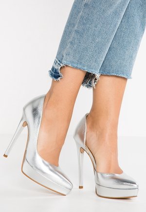 Srebrne szpilki o błyszczącym metalicznym wykończeniu, na platformie, z noskiem w szpic i obcasem typu szpilka, zestawione z podwiniętymi, przetartymi dżinsowymi spodniami.