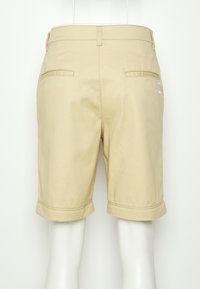 Shorts en coton beige avec des revers, deux poches arrière et un petit logo sur le côté droit. Texture lisse et coupe ajustée.