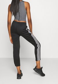 Svarta träningsbyxor med vita sidostripes och tryckknappar, ihop parat med en grå randig ärmlös topp, sedd bakifrån.