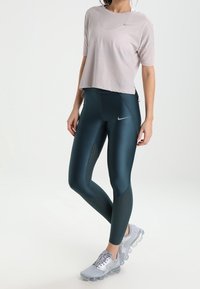Top atlético de manga corta rosa claro con textura de malla; combinado con leggings de talle alto en verde azulado oscuro con un patrón de puntos y zapatillas grises.