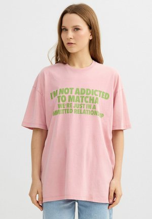 Joven con cabello largo y castaño lleva una camiseta rosa de gran tamaño que dice: "NO SOY ADICTA AL MATCHA, SOLO TENEMOS UNA RELACIÓN COMPROMETIDA."