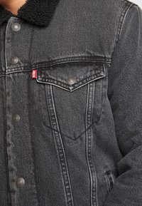 Mustat farkkutakki, jossa on nappikiinnitteinen rintatasku, musta sherpa-kaulus ja näkyvä Levi'sin punainen tappi vasemmalla puolella.