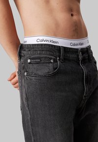 Jeans de mezclilla negros con un primer plano, presentando una cinturilla blanca con el logo de Calvin Klein y un bolsillo frontal con acentos metálicos, textura suave.