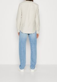 Helle beige Langarmbluse mit abgerundetem Saum, kombiniert mit lockeren hellblauen Jeans und weißen Sneakers. Die Rückansicht zeigt ein schlichtes Design.