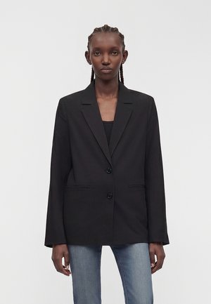 Zwarte oversized blazer met twee knopen gedragen door een vrouw met gevlochten haar, gecombineerd met medium washed blauwe spijkerbroek tegen een effen achtergrond.