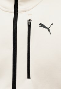 Κοντινό πλάνο κρεμ υφάσματος με μαύρο κάθετο φερμουάρ και μικρό μαύρο λογότυπο Puma στη δεξιά πλευρά της περιοχής του στήθους.