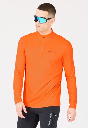 Helles orange Langarmshirt mit einem halben Reißverschluss-Kragen, elastischem Stoff und dezentem Markenlogo. Getragen mit schwarzen, eng anliegenden Hosen.