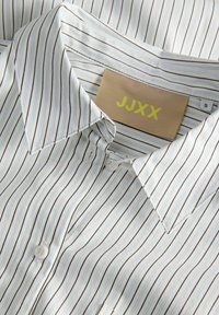 Striped button-up shirt in white and light blue, featuring thin brown stripes. The collar is pointed, with a beige label displaying "JJXX."